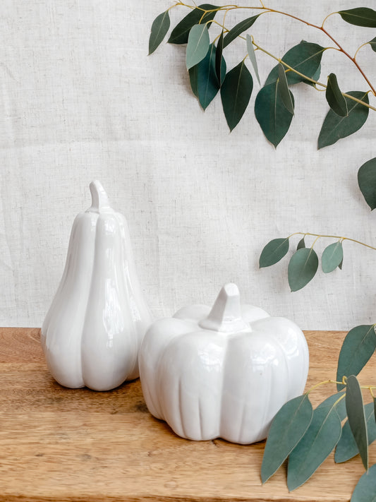 Two white ceramic gourds on a wooden surface with green leaves in the background.