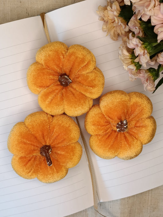 Three orange velvet pumpkins on an open book with a bouquet of flowers in the background.