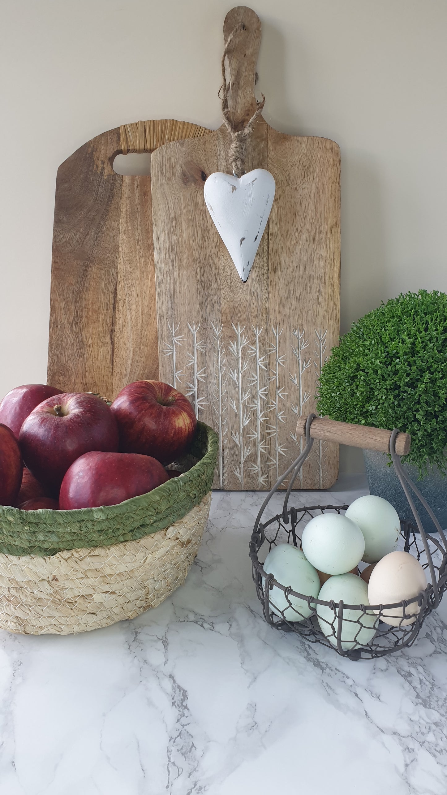 Olive trimmed straw basket with apples shown here with wooden chopping boards, egg basket and faux hebe from Little Wren Interior's range of rustic kitchen accessories