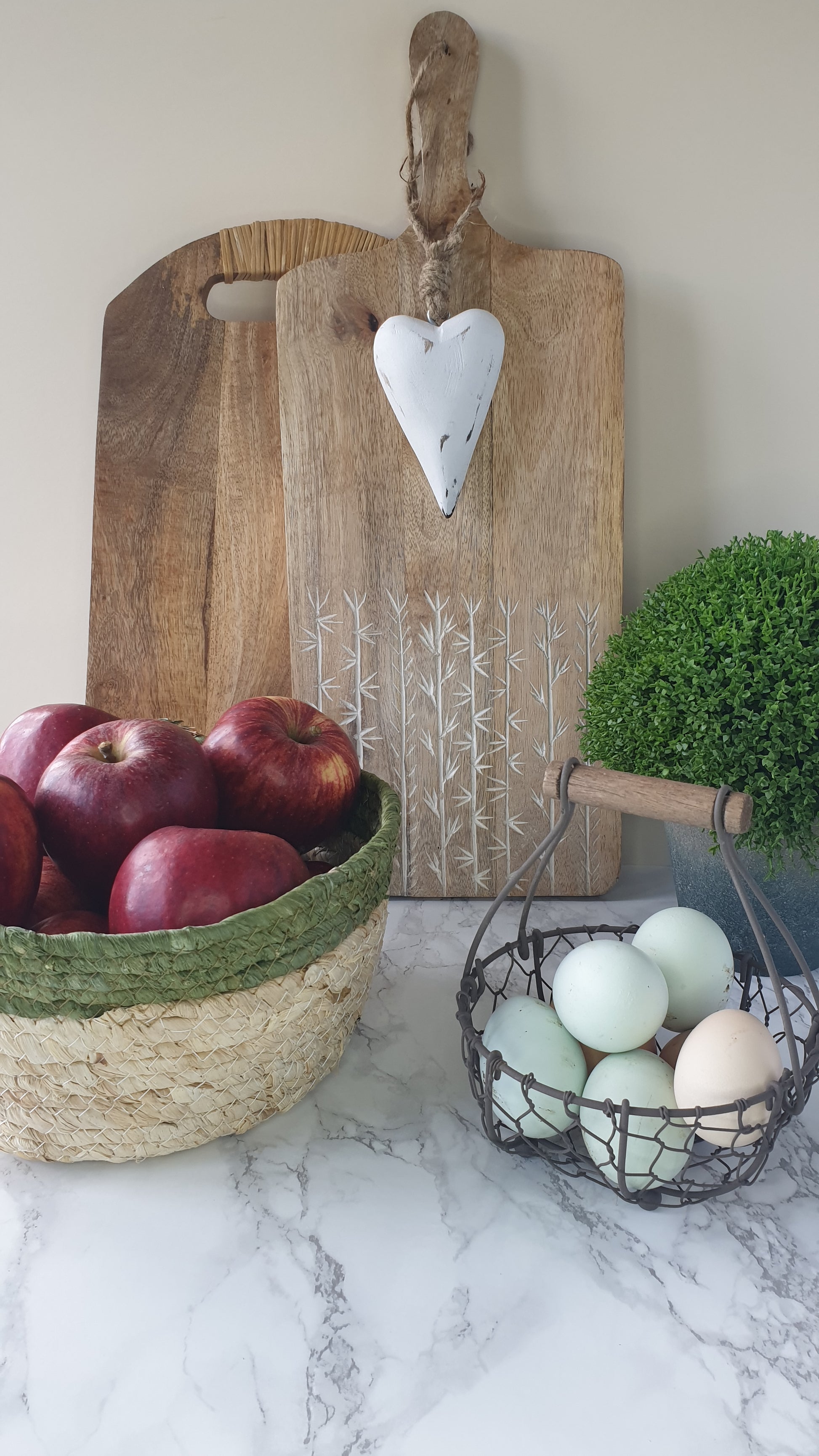 Olive trimmed straw basket with apples shown here with wooden chopping boards, egg basket and faux hebe from Little Wren Interior's range of rustic kitchen accessories