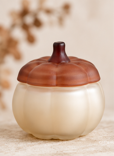A small pumpkin-shaped container with a ribbed cream body and a brown lid designed to look like a pumpkin, set against a warm beige background with soft blurred foliage.