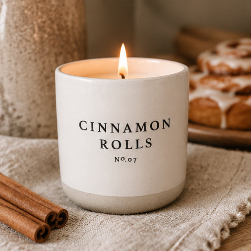A lit candle in a matte white ceramic jar with the words 'CINNAMON ROLLS' and 'No.07' printed on the front.