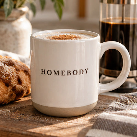 A white ceramic coffee mug with the word 'HOMEBODY' printed in black serif letters on the front, filled with a hot beverage and topped with cinnamon. The mug sits on a rustic wooden table with a pastry, a glass of coffee, and a striped cloth nearby.