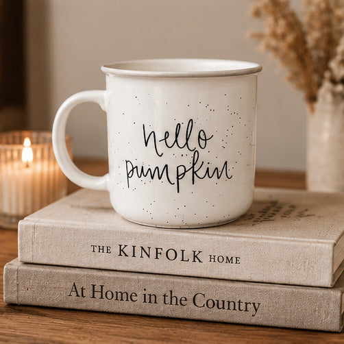An off-white speckled enamel-style mug with the phrase 'hello pumpkin' in black script sits on top of two stacked books on a wooden surface. A lit candle glows in the background on the left, and dried flowers appear blurred in the background on the right.