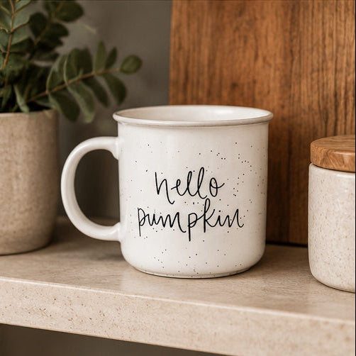 A white speckled enamel-style mug with the phrase 'hello pumpkin' in black script, sitting on a beige countertop in a kitchen setting with a plant and wooden background visible.