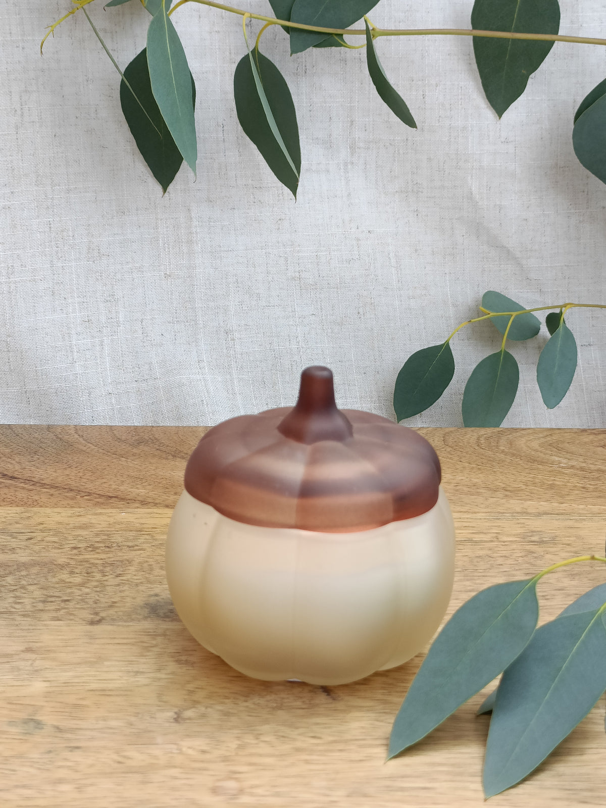 Decorative pumpkin jar candle with a brown lid on a wooden surface with green leaves.