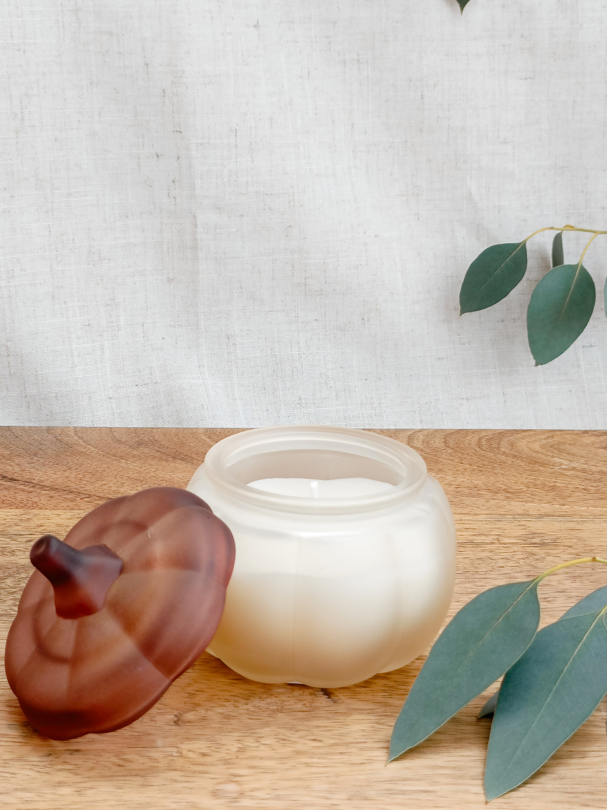 White candle in a glass jar with a brown lid designed to look like a pumpkin on a wooden surface, surrounded by green leaves.