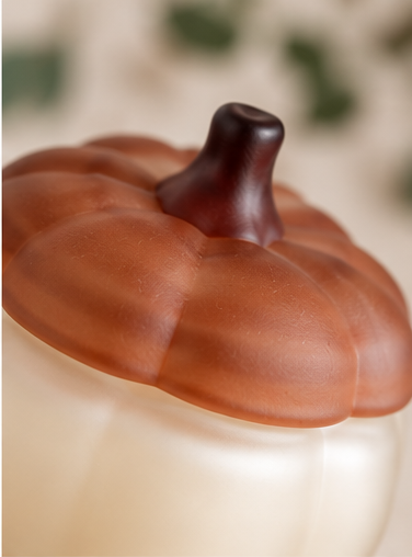 A close-up of a decorative pumpkin-shaped container or candle with a glossy brown lid and a cream-colored body, set against a softly blurred warm neutral background.