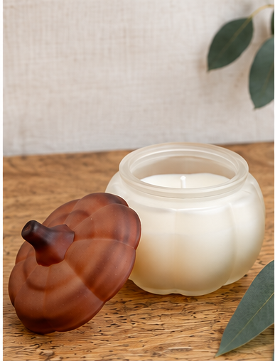 A frosted glass candle jar with a matching pumpkin-shaped lid, shown on a wooden surface with a neutral fabric backdrop and eucalyptus leaves.