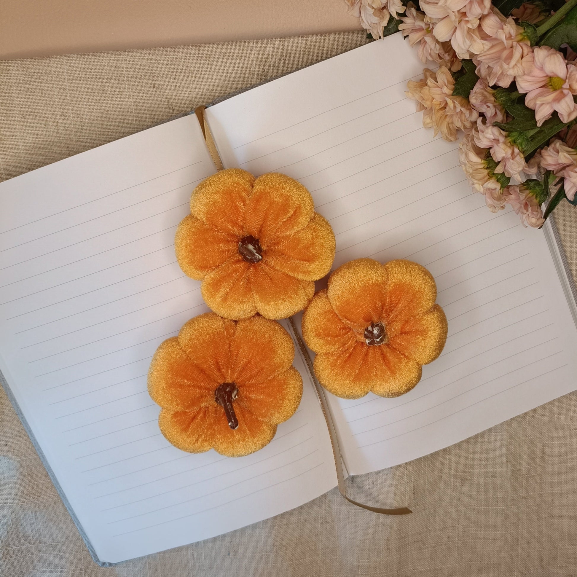 Velvet orange pumpkins on an open book with a bouquet of flowers and cinnamon sticks in the background.