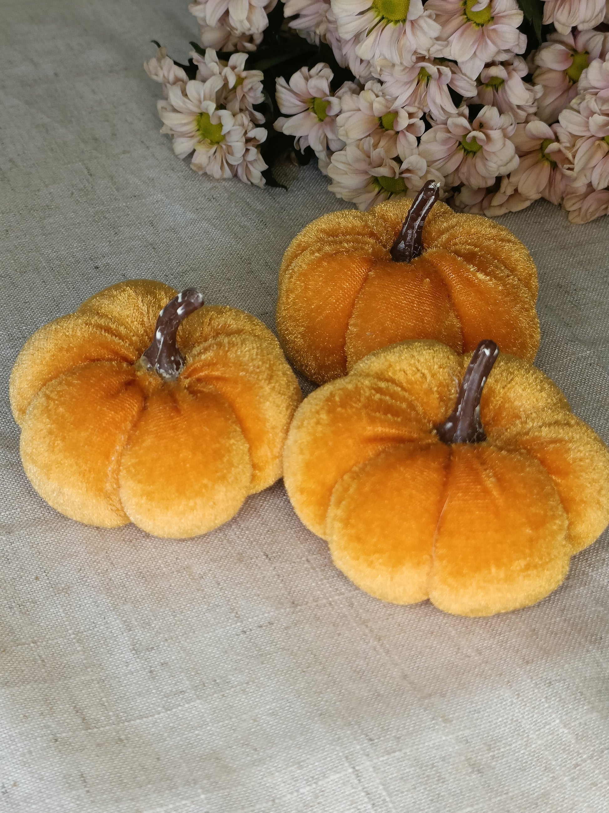 Three orange velvet pumpkins on a textured surface with flowers in the background
