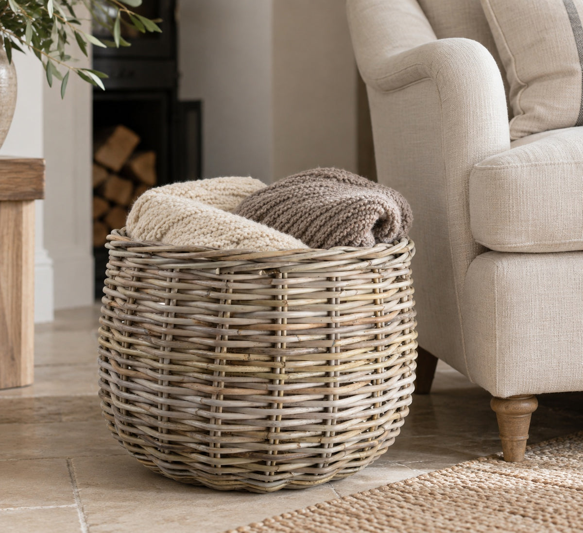 A round woven wicker basket filled with folded knit blankets, placed beside a light beige armchair in a softly lit living room.